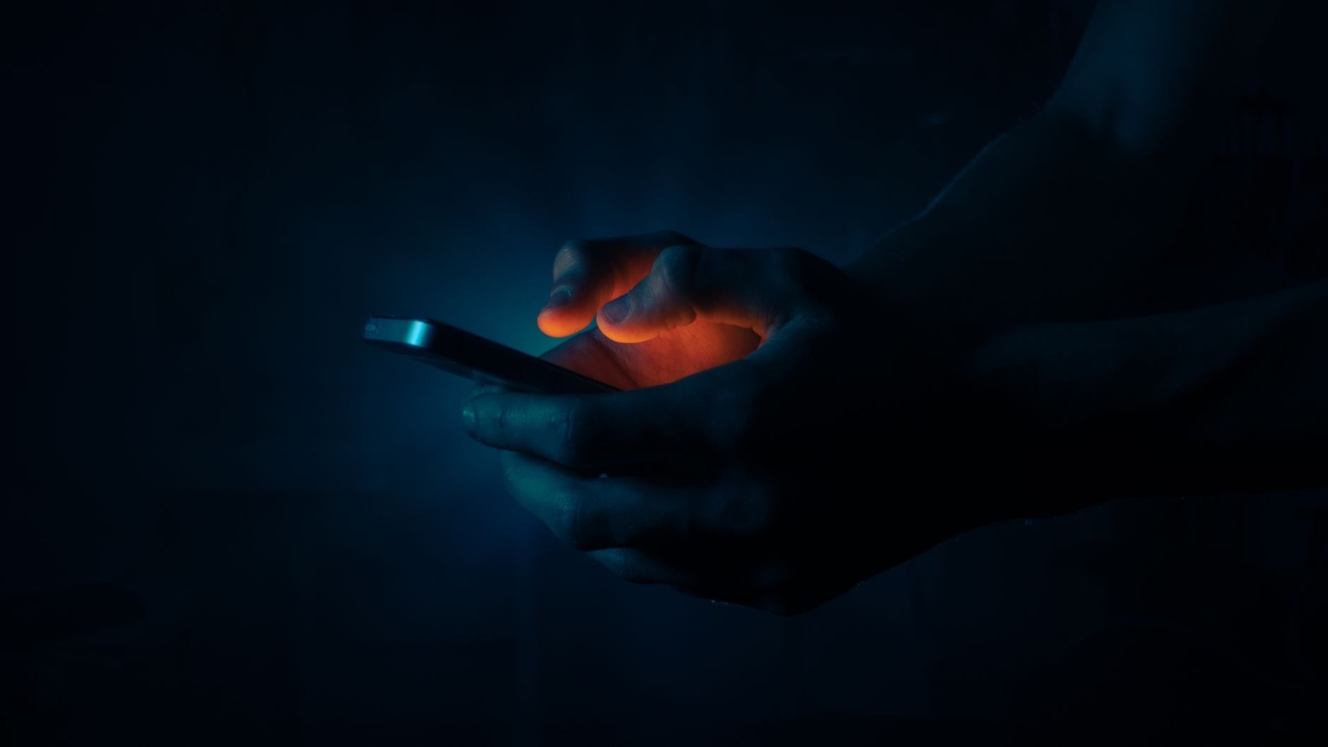 Dark and moody image of hands holding a smartphone, with a glow from the screen.