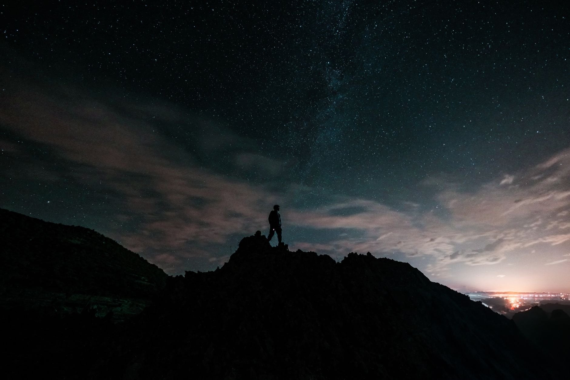 A lone hiker stands atop a mountain peak silhouetted against a starry night sky.