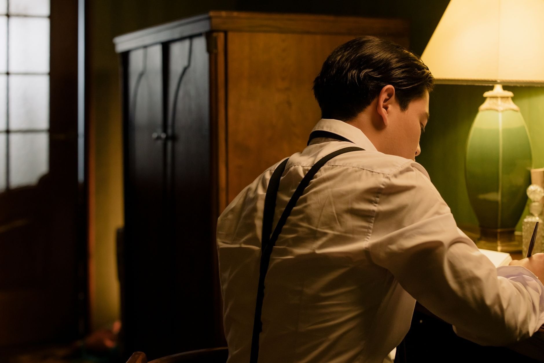 A man seated at a desk writing, illuminated by warm lamp light in a vintage style room.
